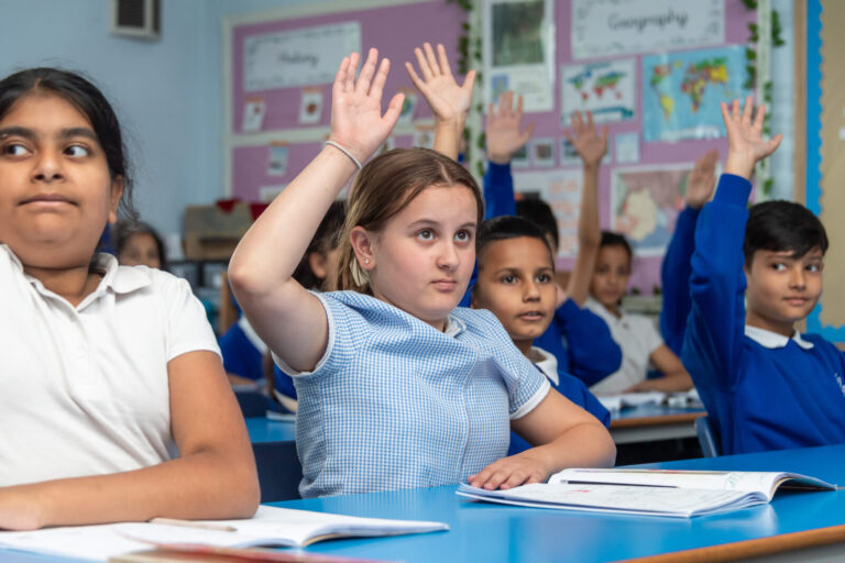School children at desks holding up there hand to answer question